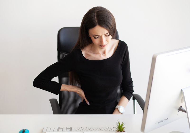Una chica sentada en la oficina con la mano en la lumbar porque tiene dolor de espalda.